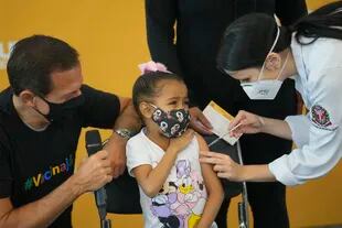 Una trabajadora de la salud aplica la vacuna COVID-19 de Pfizer a Valentina Moreira, de 6 años de edad, mientras el gobernador de Sao Paulo, Joao Doria, está junto a ella con un micrófono el viernes 14 de enero de 2022, en el Hospital da Clinicas en Sao Paulo, Brasil. (AP Foto/Andre Penner)