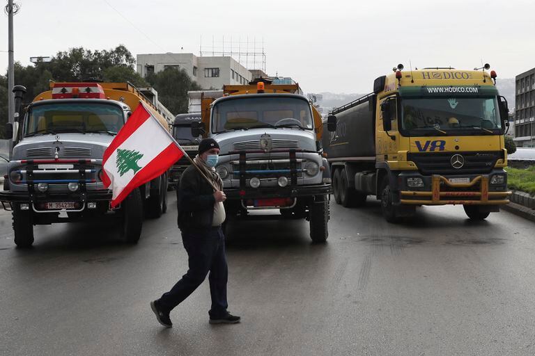 Un conductor de camiones alza una bandera libanesa mientras otros bloquean una autopista durante una huelga general de los sindicatos del transporte y trabajadores que paralizó el Líbano en protesta por el deterioro de la situación económica, en Beirut, jueves 13 de enero de 2022. (AP Foto/Bilal Hussein)