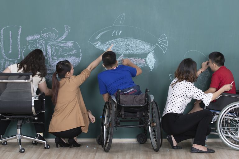 Group of special students in classroom, a down syndrome girl, two handicapped boys and  two female teachers drawing and painting on black board together.