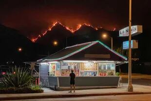 Un hombre se encuentra frente a un restaurante de comida para llevar mientras las llamas arden en las colinas del Bosque Nacional de Ángeles en el fondo en medio de los incendios forestales en California en septiembre de 2020.