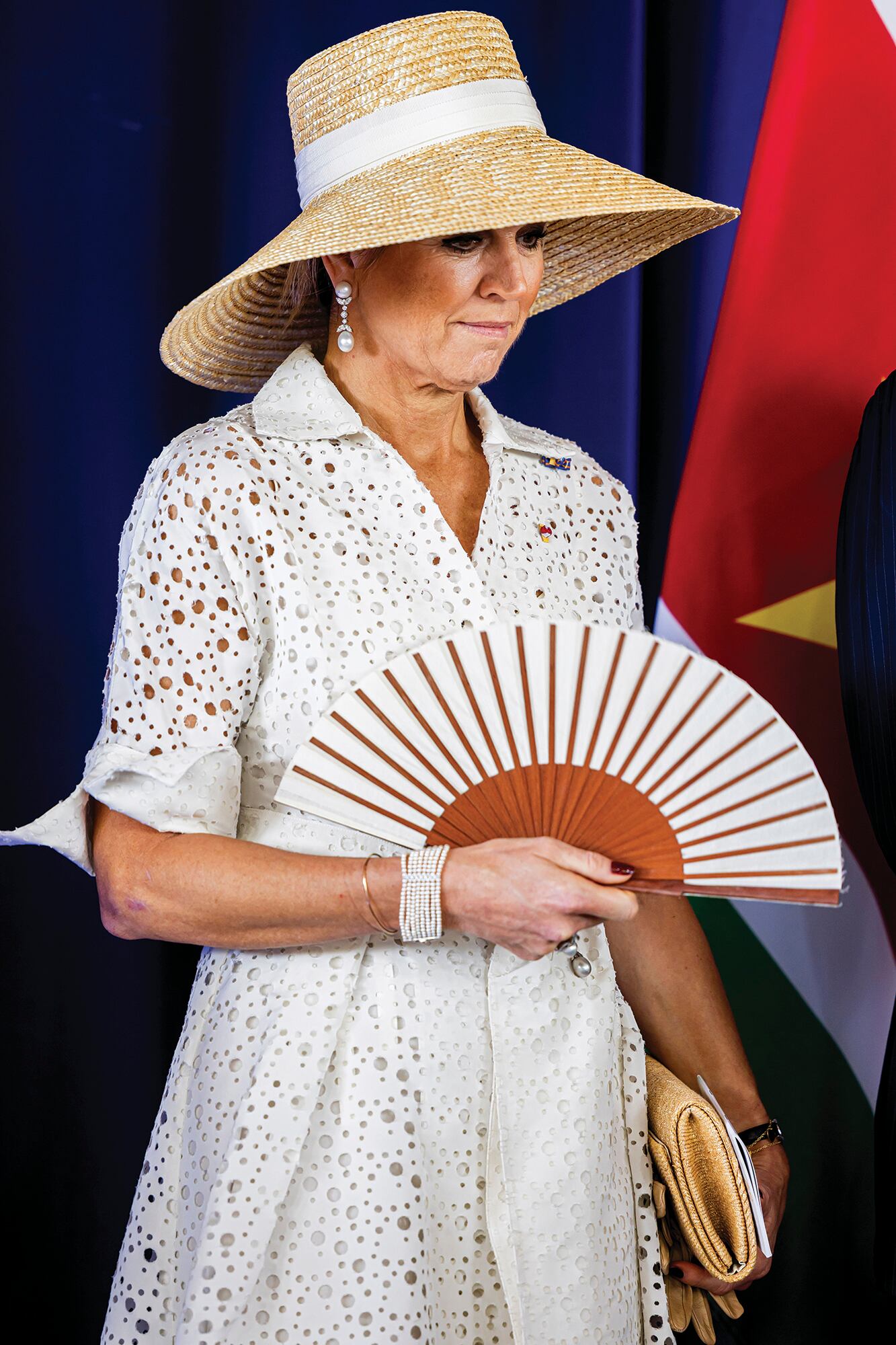 Para la ceremonia de bienvenida, la Reina volvió a elegir a Natan, esta vez con un estreno: un vestido calado y cruzado de puños anchos y lazo en la cintura, rematado por un broche de perlas y diamantes. Los accesorios –pendientes, anillo y brazalete de varias vueltas– también son de perlas. Completó el look con una gran pamela de rafia