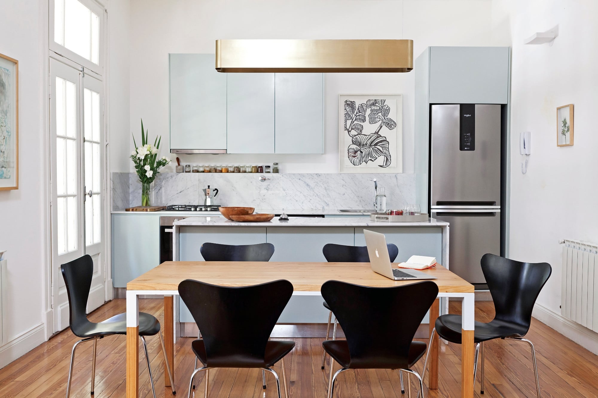 Las sillas negras estilo Jacobsen ayudan a la transición entre el living y la cocina. Sobre la mesa (Números Primos), la estilizada lámpara colgante de bronce (La Feliz) conversa con el cuadro de María Marta Fasoli (Izq.), enmarcado en madera al natural.