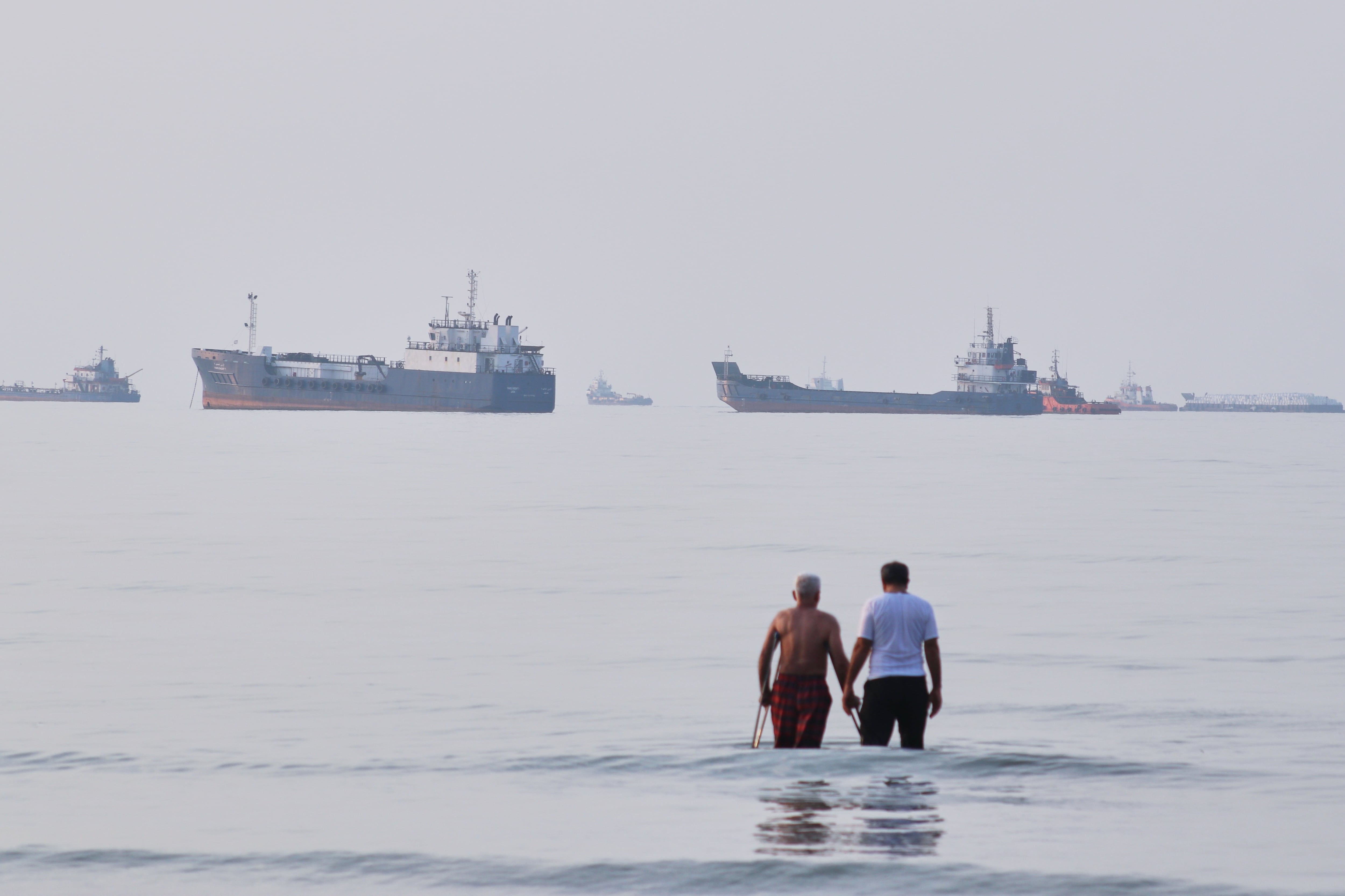 Dos hombres caminan en las aguas del estrecho de Ormuz con barcos fondeados más lejos, cerca de Bandar Abbas, Irán