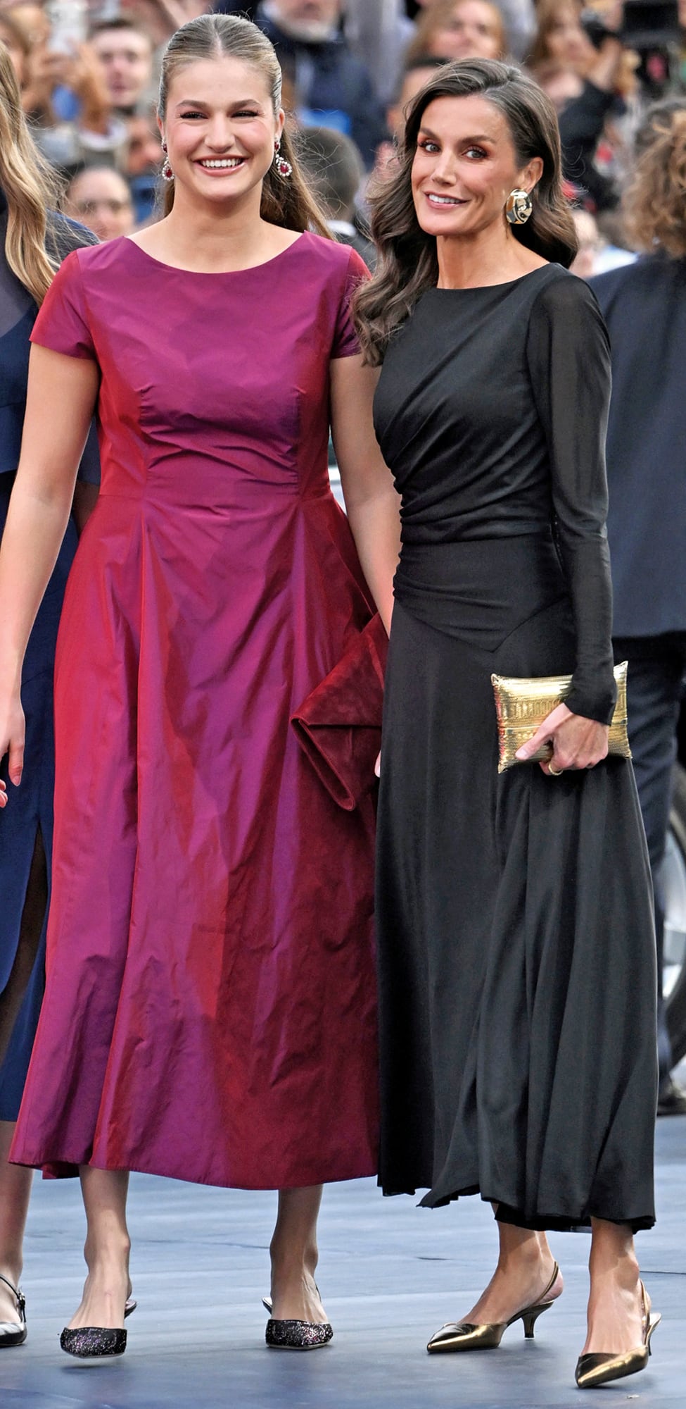 Madre e hija, impactantes. La princesa Leonor con un vestido bordó firmado por Hannibal Laguna White, de escote bote, cuerpo ceñido y falda midi en línea “A”, potenciado con los pendientes de diamantes y rubíes de Van Cleef & Arpels del cofre de su abuela. Lo complementó con clutch a tono de Magrit y zapatos Carolina Herrera. Letizia, en cambio, fue en total black, con un vestido sostenible (confeccionado con un material que se obtiene de combinar tejidos recuperados y pulpa de madera, desarrollado por la empresa I+D textil PYRATEX) entallado y drapeado, que combinó con sobre dorado de Begüm Khan, zapatos de Magrit y grandes aros
