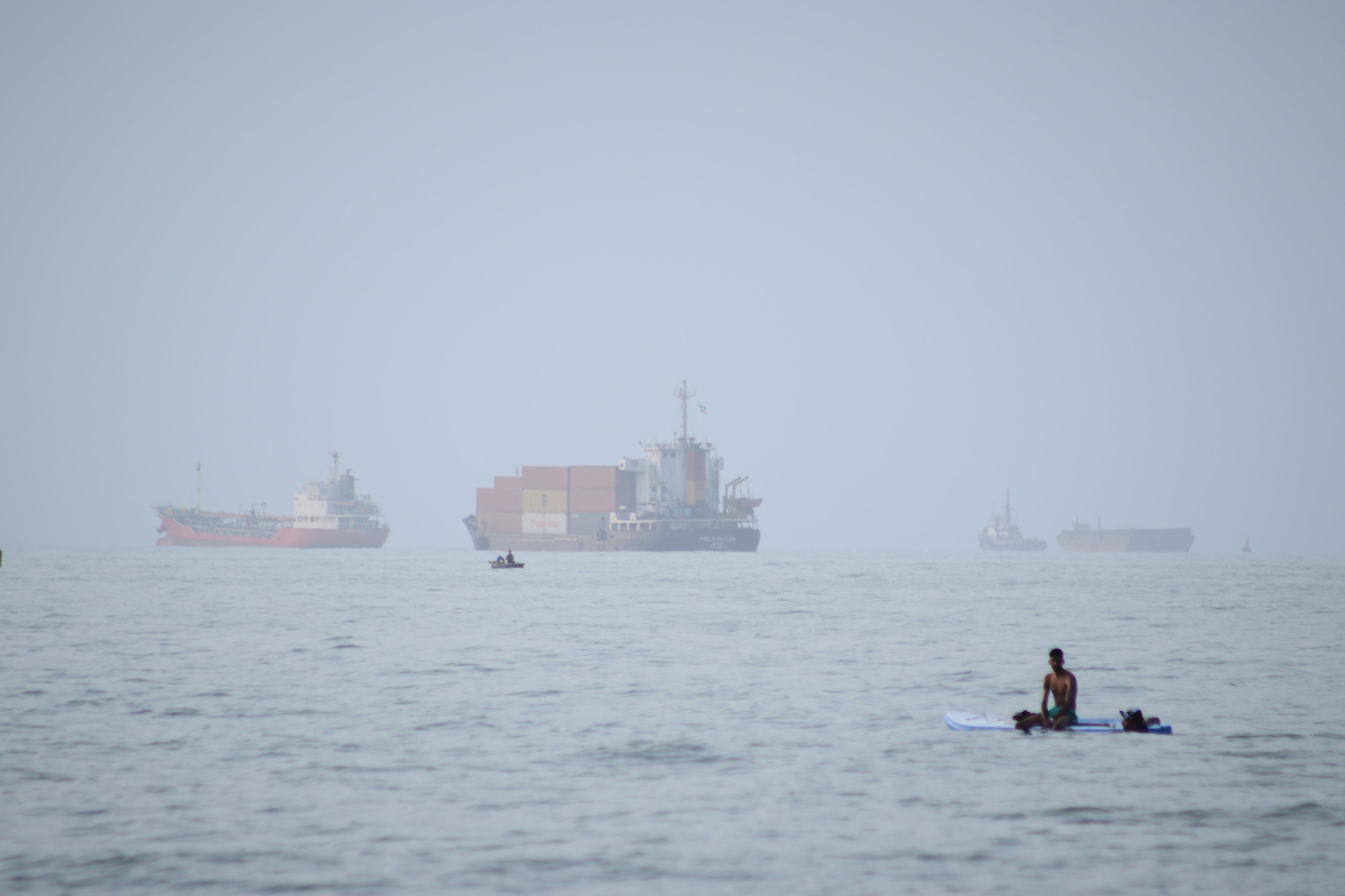 Un hombre se sienta en una tabla de surf con un barco portacontenedores al fondo junto a otros buques comerciales, en el estrecho de Ormuz, desde Bandar Abbas, irán