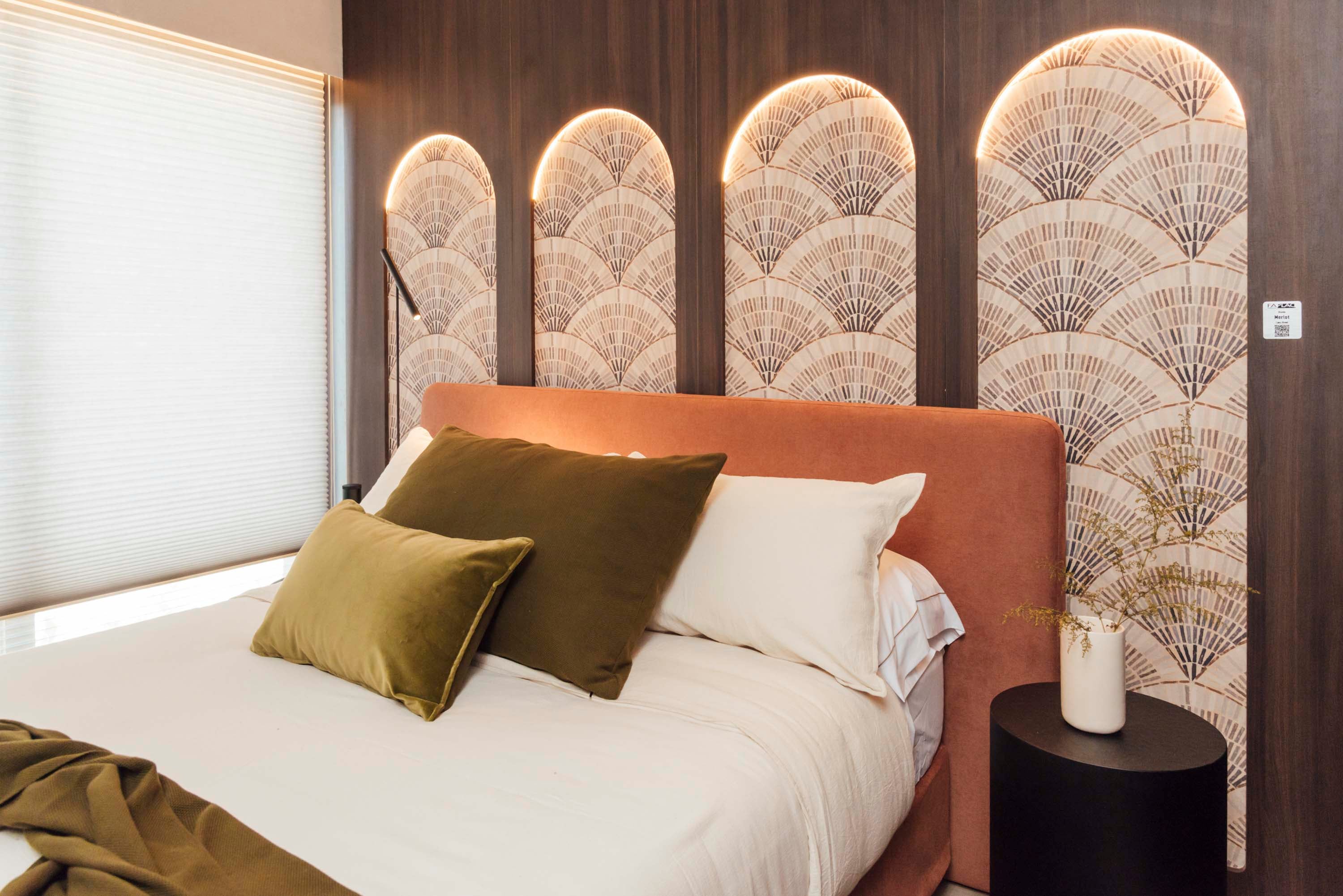 En el dormitorio del espacio de Halesa, la pared revestida con melamina Merlot (Faplac) tiene arcos calados retroiluminados que dejan ver un empapelado con motivos de abanicos (Tintachini, aplicado por Empapelarte). Cabecera de cama en terciopelo terracota y almohadones en tonos oliva.
