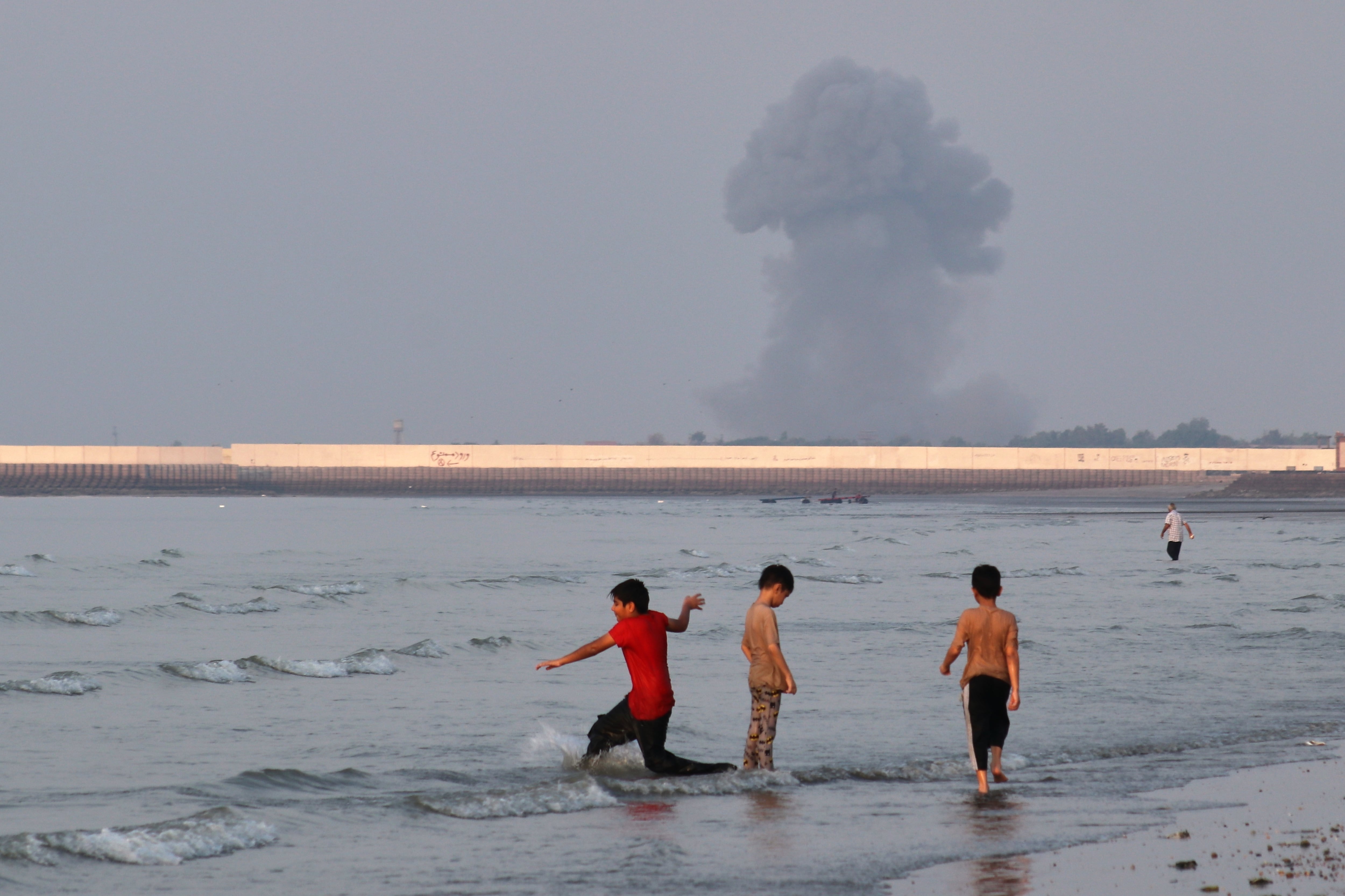 Tres niños juegan en el agua en el estrecho de Ormuz mientras al fondo se alza una columna de humo de una explosión, cerca de Bandar Abbas, Irán, el lunes 13 de julio de 2026 (Razieh Poudat/ISNA via AP)