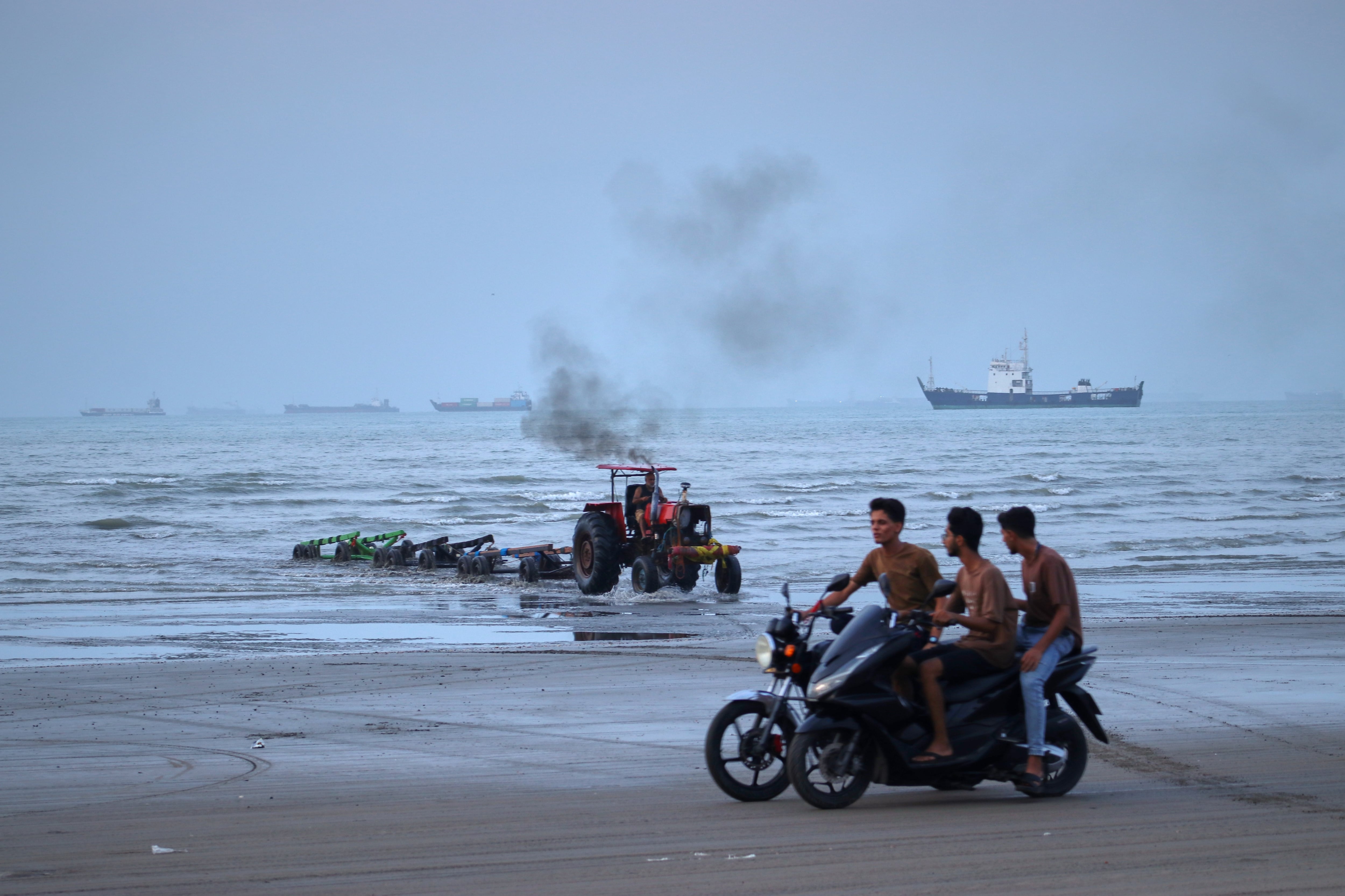 Jóvenes a bordo de motocicletas a lo largo de la orilla mientras un tractor saca equipo del agua y al fondo se pueden ver buques mercantes aparentemente anclados en el estrecho de Ormuz