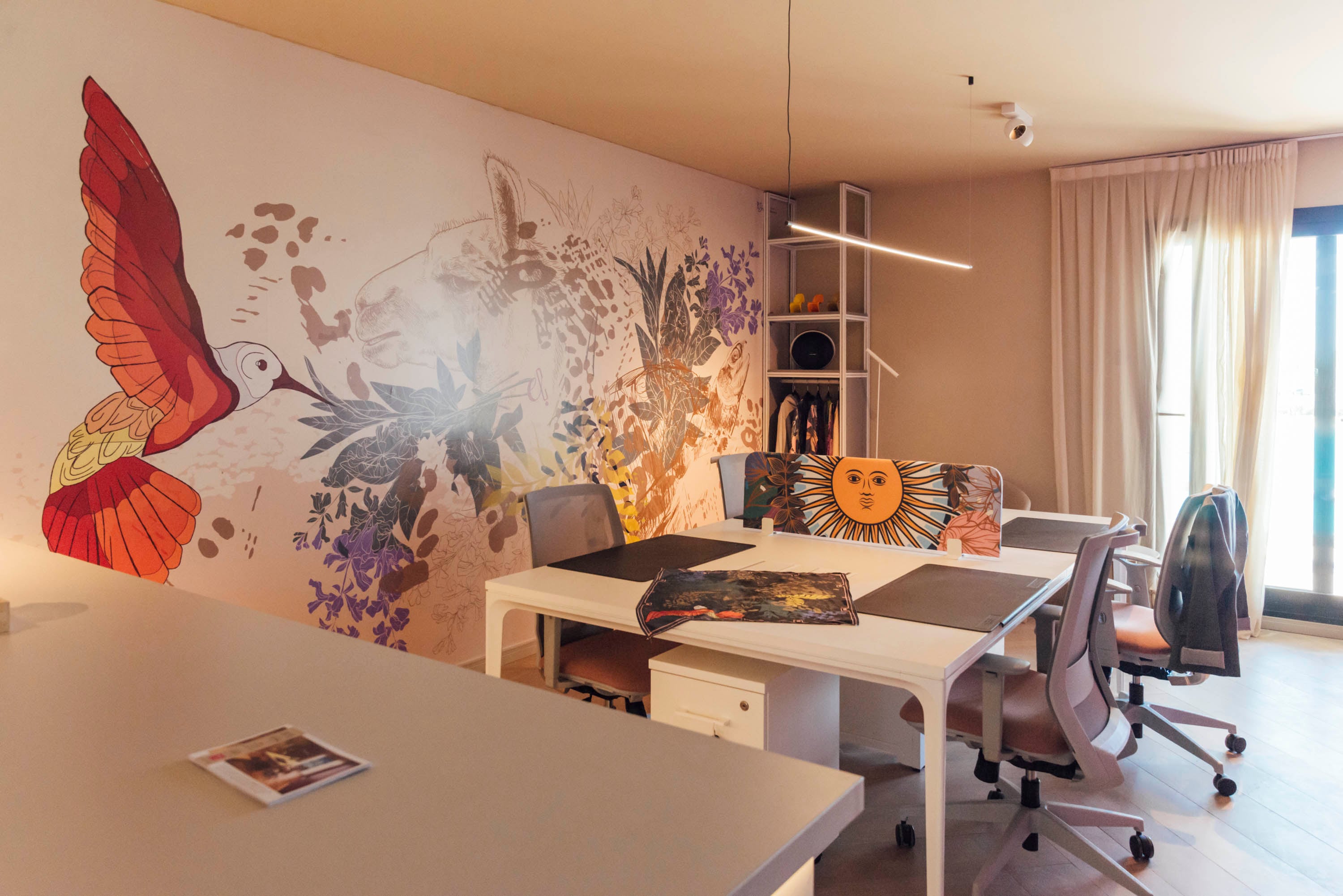 Interior del espacio de trabajo que es el showroom de Era Carlota con un gran mural con colibrí, fauna y flora sobre la pared principal. Escritorios blancos y sillas ergonómicas en tonos rosados de Grupo(a)2. Iluminación lineal suspendida.