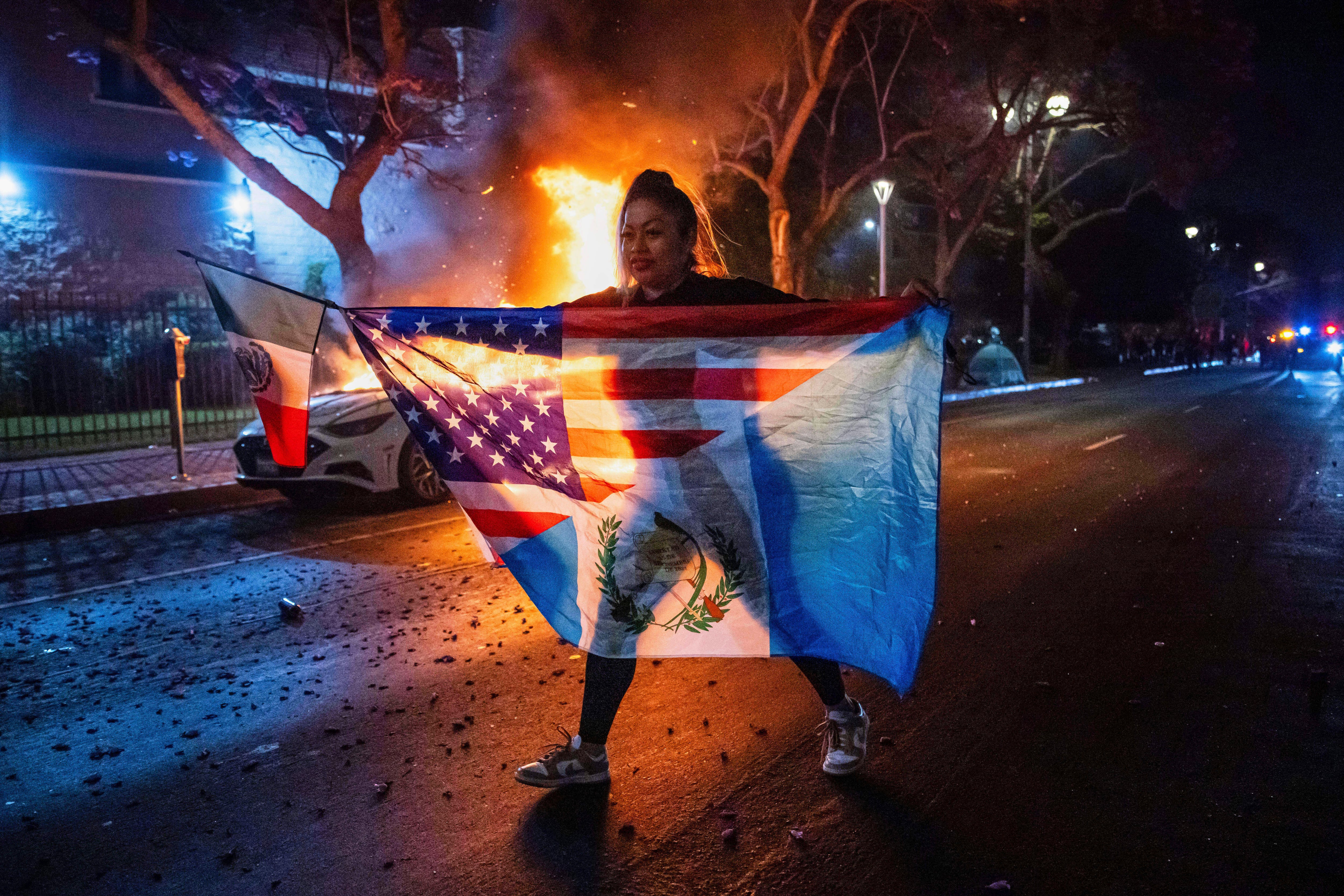 Una persona con una combinación de banderas camina frente a un auto incendiado durante las protestas por las redadas del gobierno de Trump en Los Ángeles, el lunes 9 de junio de 2025. (AP Foto/Ethan Swope)