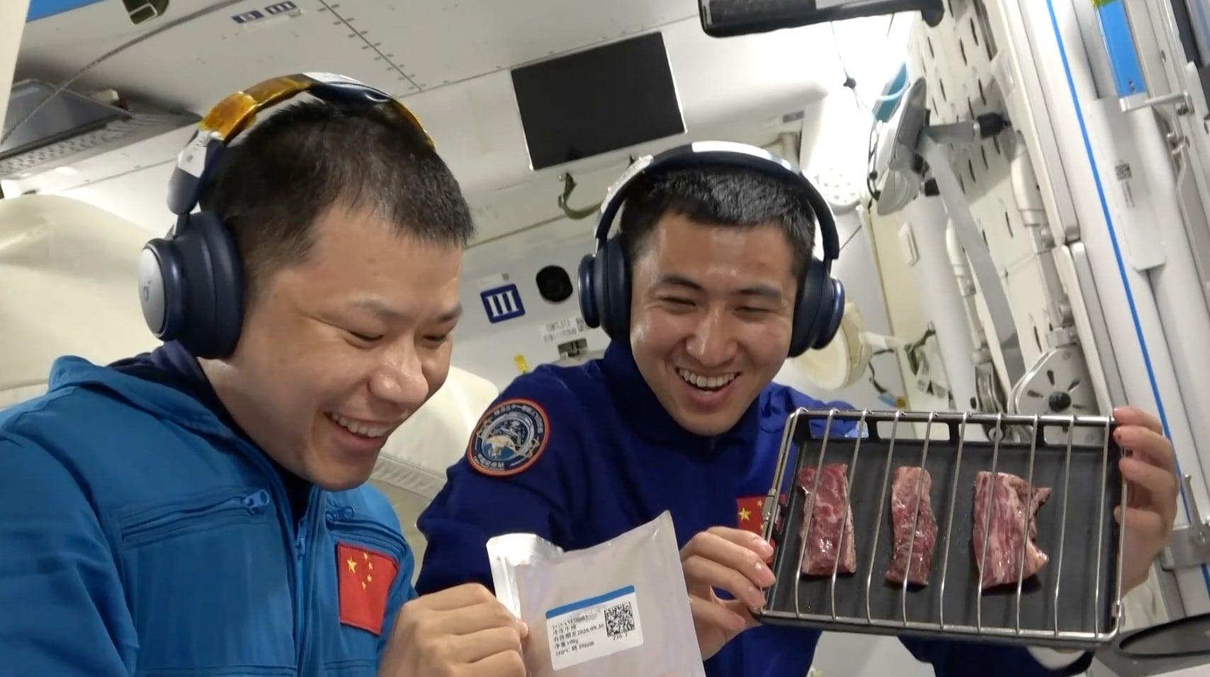 El miembro de la tripulación de Shenzhou 20, el coronel Wang Jie (a la izquierda), y el ingeniero de vuelo espacial de Shenzhou 21, el mayor Wu Fei, se preparan para asar un plato de carne con pimienta negra en un horno especialmente construido, entregado este martes por la nave espacial Shenzhou 21 a la estación espacial Tiangong.