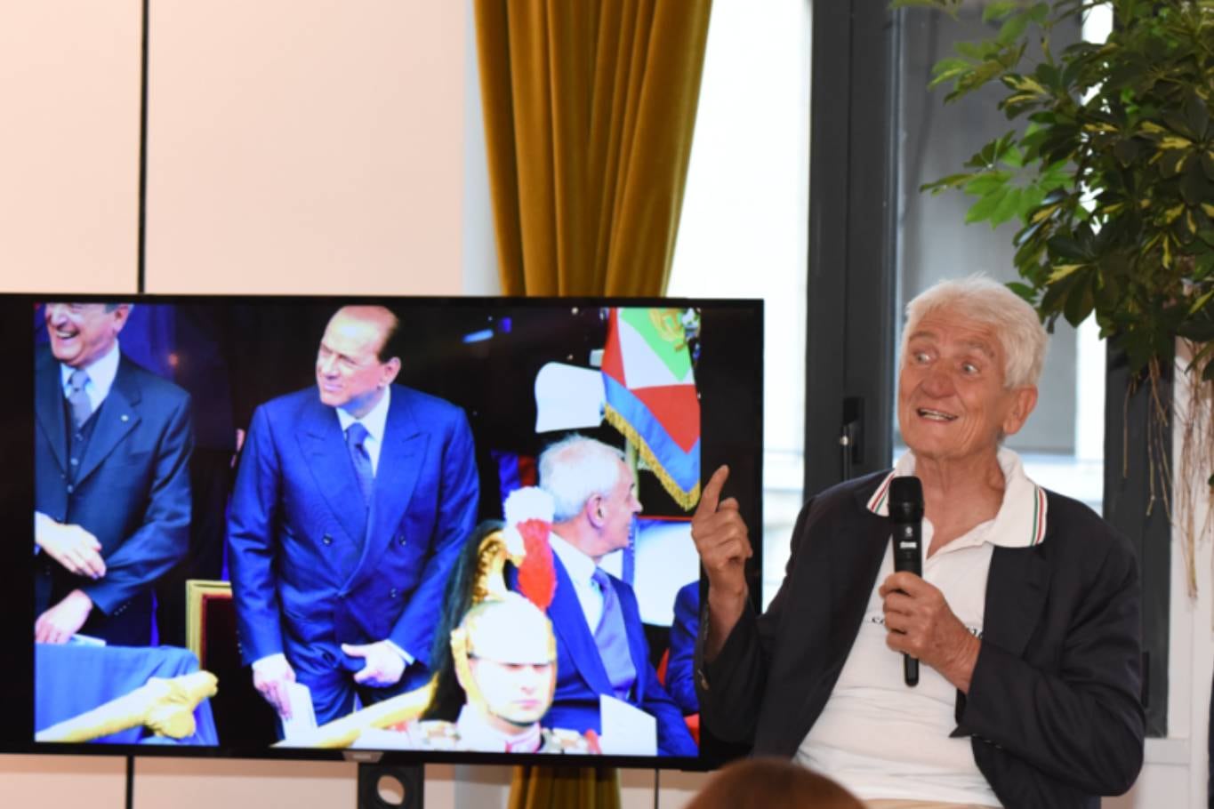 Umberto Pizzi en una conferencia con una de sus obras detrás, la de Silvio Berlusconi tomándose sus partes, una foto que ningún medio quiso comprar