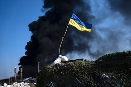 Una bandera ucraniana se ve en la parte superior de un puesto de control mientras el humo negro se eleva desde un almacenamiento de combustible del ejército ucraniano luego de un ataque ruso, en las afueras de Kiev, Ucrania