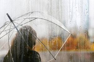 A woman silhouette with transparent umbrella through wet window with drops of rain. Autumn