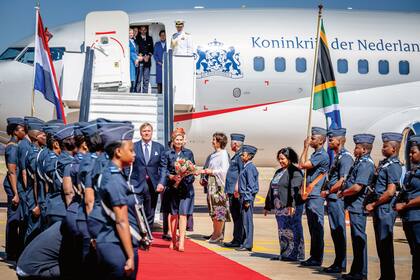 Al bajar del avión en Pretoria, la Reina llevaba un vestido camisero azul marino, creación de Jan Taminiau, que ya lució en otras ocasiones. Esta vez, lo acompañó con accesorios en naranja –el color de la casa Orange– que combinaban a la perfección: tocado de Fabienne Delvigne, clutch Banana Republic y stilettos by Natan. El rey Guillermo Alejandro, en cromática sintonía con su mujer, optó por un traje azul.