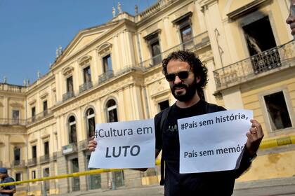 Un hombre en la puerta del museo sostiene dos carteles con leyendas: "Cultura en duelo", izquierda, y "Un país sin historia es un país sin memoria"