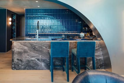 Barra-cocina del espacio "Vértice" con gran arco estructural (Artefacto BA) que marca el acceso a la cocina, revestida integralmente en cerámicos Flow Cosmic Blue (San Lorenzo Design). Mesada de Cuarcita Matarazzo (De Stefano 1913, ejecutada por Artecnia). Banquetas altas tapizadas en terciopelo azul.