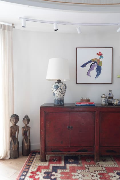 Buffet rojo, lámpara de mesa, figuras apoyadas en el piso y kilim ‘Uzbal’ de lana teñida con pigmentos naturales, originario de Uzbekistán (todo de Desde Asia). Obra ‘SerN20’, de Sol Rubino (Galería Pared).