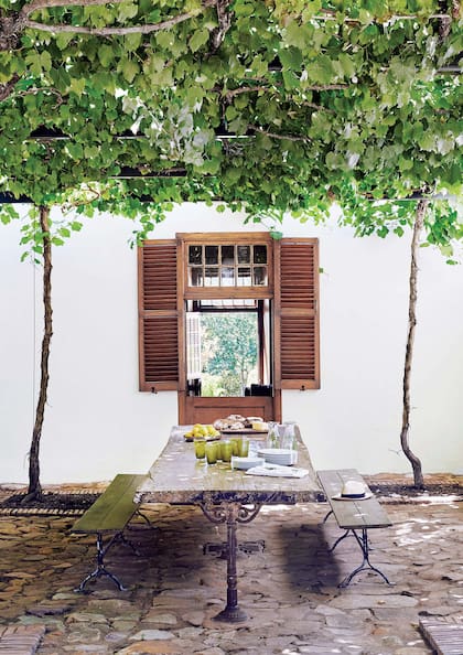 Clásico que nunca pasa de moda, en esta finca en Sudáfrica la parra y los pisos de piedra se conjugan con austeridad y elegancia. Siguiendo la línea, un juego de mesa y bancos con pies de hierro y tablones rústicos de madera ofrece un rincón perfecto para tomar algo fresco o comer en verano