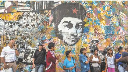 Consumidores hacen fila para comprar alimentos en Caracas bajo la mirada de un Simón Bolívar con boina.