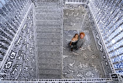 Cox y su esposa posan con la gran escalera de la casa donde se ve de piso a techo la obra que no deja un espacio libre