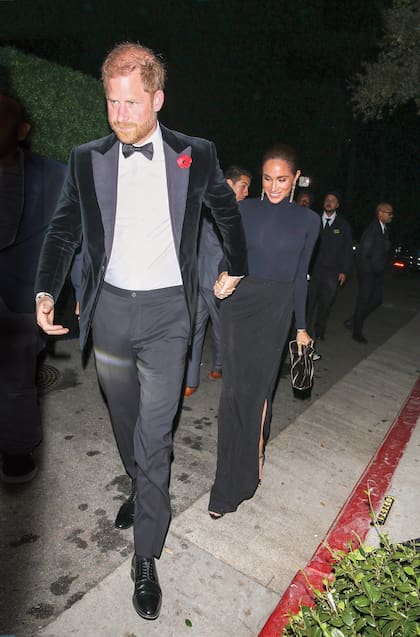 De impecable smoking, el príncipe Harry llega a la fiesta junto con Meghan Markle, con un top de cuello alto y mangas largas y falda drapeada a la altura de la cintura. El clutch es de Aquazzura