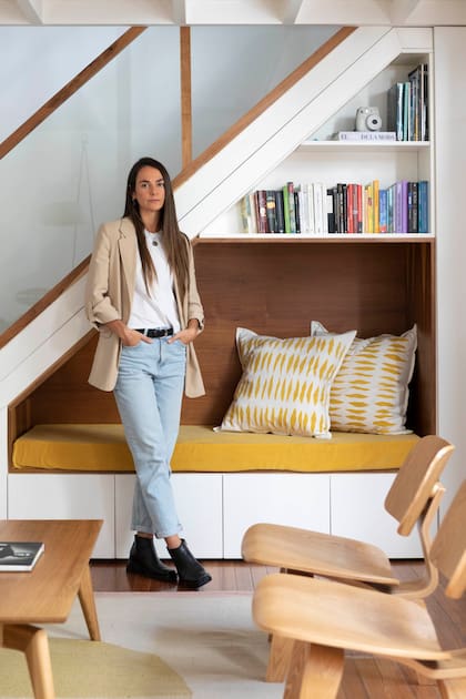 Delfina Carbó en el living, espacio que refleja su “diseño consciente”. El lugar bajo la escalera cobró propósito (Estudio DC por Carpintería Gabriel Veiras) mediante una estructura ad hoc con cajones inferiores, un banco con colchoneta y almohadones (Mercedes Segura), además de estantes para libros.