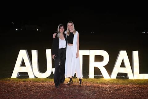 Delfina Lanusse y Chantal Leclercq posan juntas al aire libre, de noche, frente a un cartel luminoso de la Universidad Austral donde estudiaron juntas y se hicieron amigas íntimas