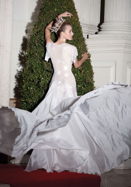 Diseño de Gino Bogani con la parte superior en encaje bordado con flores de satén, mostacillas y perlas, y falda de organza de seda natural con vuelo. Tocado de perlas y cristales y pendientes con filigrana, cristales y strass