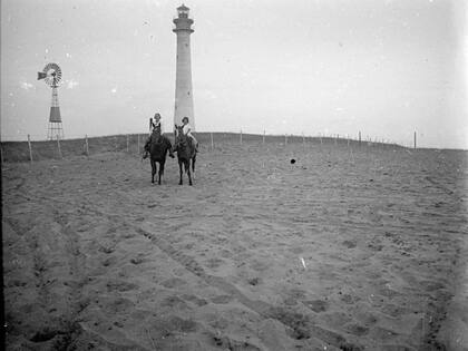 Dos personas cabalgan con el faro de Quequén en el fondo. El nombre de Quequén proviene de ‘Kem Kem’, del idioma de los pueblos originarios y significa "barrancas altas".