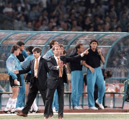 El banco de suplentes de la selección argentina en Italia 90. El DT Bilardo dando indicaciones a los jugadores que están en el campo de juego. Detrás de Bilardo está su asistente Pachamé y el doctor Madero atendiendo a un jugador lesionado. 