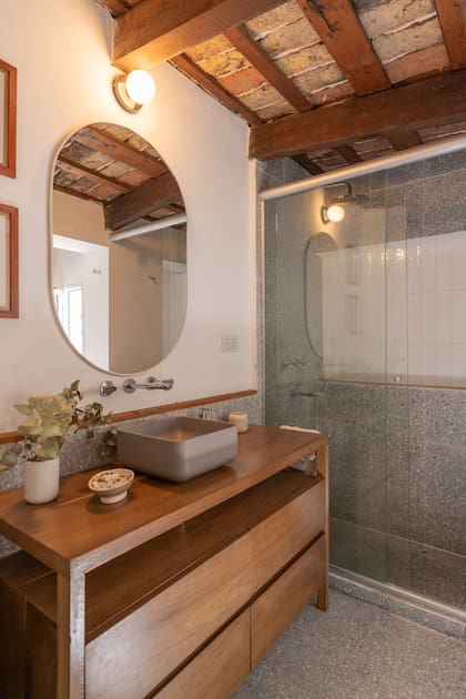 El baño combina materiales cálidos y texturas originales: techo de ladrillo visto con vigas de madera, mesada en madera y detalles contemporáneos que dialogan con la historia del espacio.