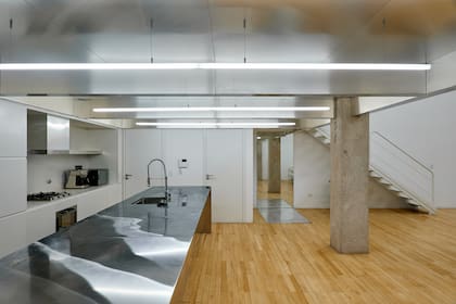 El cielorraso de la cocina mantiene el mismo ritmo de vigas que en el resto del ambiente. Lámparas de 3 metros (Huup). Mueble con nicho y mesada de Neolith. La mesada de la isla es de acero.