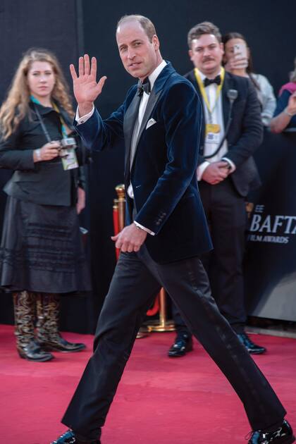 El hijo del rey Carlos III saluda al público en la red carpet de los BAFTA. Llevó un saco de terciopelo azul marino con solapas negras, combinado con pantalones negros, moño a tono y camisa blanca.
