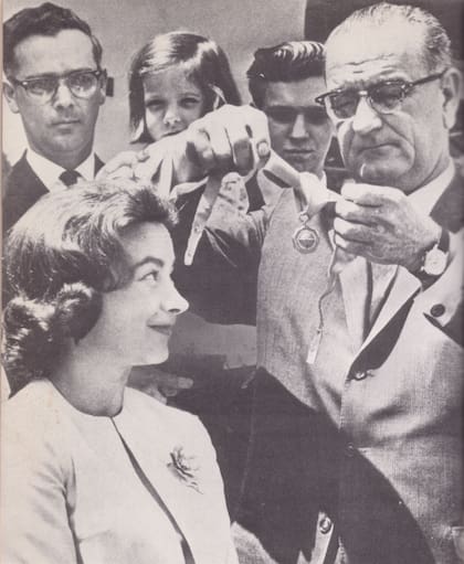 El presidente Lyndon B. Johnson condecora a Jerrie Mock en el Rose Garden de la Casa Blanca, el 4 de mayo de 1964. Detrás de la aviadora, observan la escena con mucha emoción su marido, Russell Mock, y sus hijos, Valerie y Gary. La medalla de oro, otorgada por la Agencia Federal de Aviación, es la máxima condecoración para un aviador civil en los Estados Unidos.