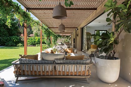 El techo de la galería tiene listones de madera que dejan pasar la luz natural. En primer plano, vemos el sector del living con sillones de respaldo circular y una mesa baja con tapa de mármol. Más atrás, el comedor con muebles macizos de madera. La parrilla está acompañada por una barra.