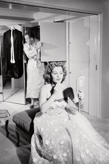 Ella se maquilla con un espejo de mano mientras su tercer marido, el actor George Sanders, la mira por el espejo. Zsa Zsa lleva puesto un strapless y el smoking de él todavía está colgado. Era el año 1950.