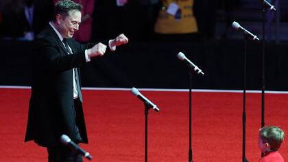 Elon Musk con su hijo, X, en el escenario del mitin de la victoria de Trump en Washington, DC. Musk sonríe y levanta las manos. La cara de X no es visible.