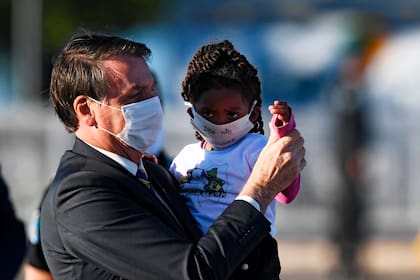 En esta foto de archivo tomada el 12 de mayo de 2020, el presidente brasileño, Jair Bolsonaro, sostiene a una niña en sus brazos, ambos con barbijos, durante la ceremonia de izamiento de la bandera antes de una reunión ministerial en el Palacio Alvorada en Brasilia, en medio de la nueva pandemia de