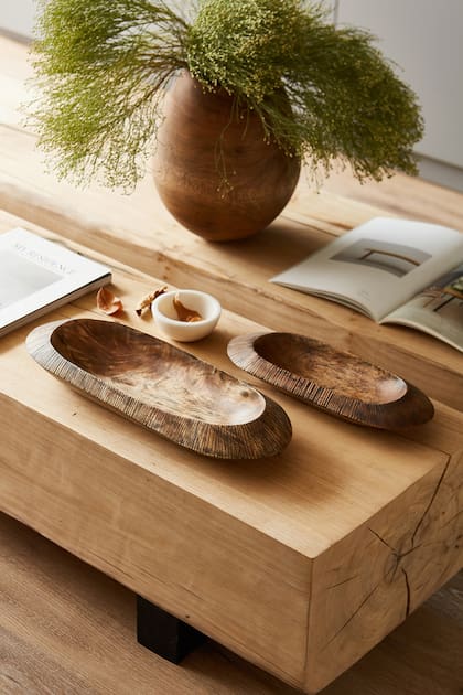 En la mesa ratona, el follaje no es tropical sino más invernal. Momento perfecto para elegir un florero de madera sin agua y bandejas talladas (Dukaan Home).
