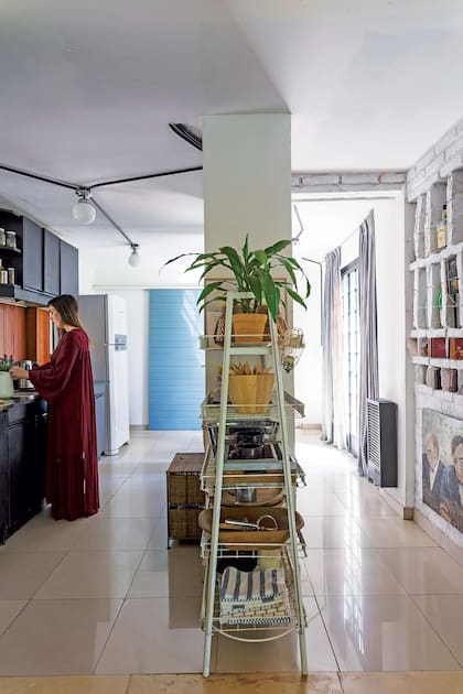 En línea con la columna estructural que divide la cocina, una estantería antigua con finos parantes blancos y base enrejada para sumar guardado.
