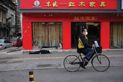 En Wuhan, un hombre pasea en su bicicleta; detrás, tirado en la calle, una víctima fatal del virus