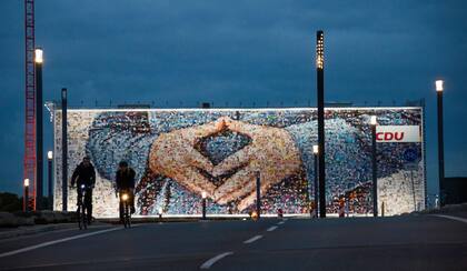 Esta foto de archivo tomada el 18 de septiembre de 2013 muestra a personas en bicicleta frente a un cartel electoral gigante con el famoso gesto con las manos en forma de diamante de la canciller alemana Angela Merkel que se exhibió en el sitio de construcción de un hotel en Berlín