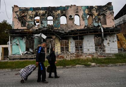 Esta fotografía, tomada el 11 de septiembre de 2022, muestra a una pareja que lleva maletas mientras camina frente a un edificio destruido en Izyum, región de Kharkiv, este de Ucrania.