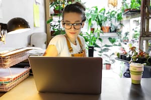 European female gardener in glasses using laptop, scrolling through social networks, reads news, coffee/tea mug on table, home garden/greenhouse on background. Cozy workplace, remote work