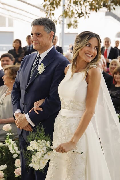 Fabián Zitta vistió a los novios. Belén lució un vestido en mikado de seda italiano, con corsage a la caja y escote bote, falda sirena con entalle y sobrefalda en capas con flores bordadas a mano con perlas y canutillos. Sobre sus hombros, llevó una capa de organza de seda de largo catedral. Lo complementó con pines para el pelo (Laura Noetinger) y zapatos en satén (Ricky Sarkany). Jorge, en tanto, estaba impecable con un traje de corte italiano confeccionado en lana
super 140 de Loro Piana.