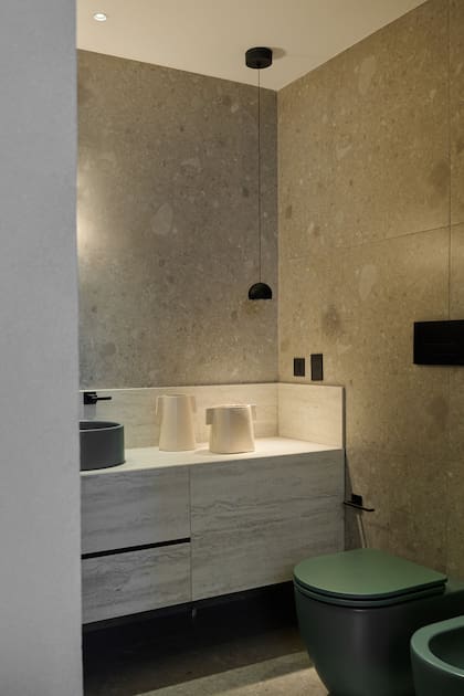 Bacha circular y sanitarios en verde oliva (Ferrum). Grifería en negro (FV). Mueble en Purastone Prima 'Travertino Navona' (De Stefano) por marmolería United Stone.