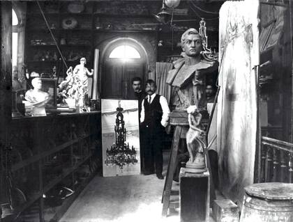 Gabriel Simonnet Dubois enseña una fotografía de la Araña del Centenario, con probabilidad en el taller de Azaretto. La maqueta del mismo artefacto se observa colgada detrás del busto de San Martín. Circa 1913.