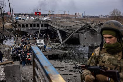 A Ukrainian serviceman looks on as evacuees cross a destroyed bridge as they flee the city of Irpin, northwest of Kyiv, on March 7, 2022. - Ukraine dismissed Moscow's offer to set up humanitarian corridors from several bombarded cities on Monday after it emerged some routes would lead refugees into Russia or Belarus. The Russian proposal of safe passage from Kharkiv, Kyiv, Mariupol and Sumy had come after terrified Ukrainian civilians came under fire in previous ceasefire attempts. (Photo by Dimitar DILKOFF / AFP)