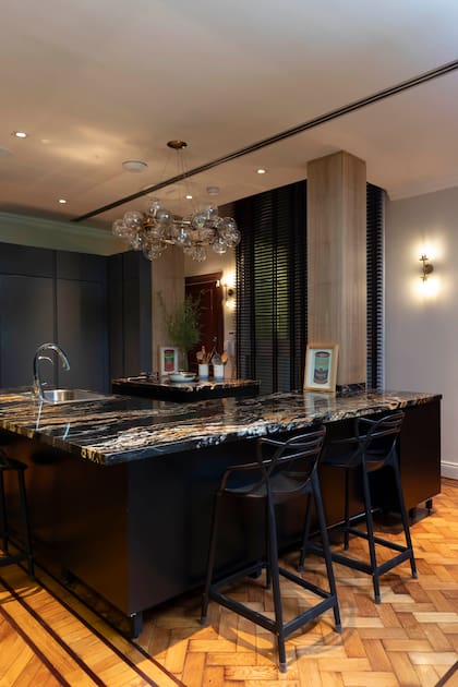 Isla protagonista con tapa de mármol veteado en tonos oscuros, mobiliario negro y piso de pinotea en espiga. La iluminación colgante aporta un toque escultórico que realza la elegancia del espacio.